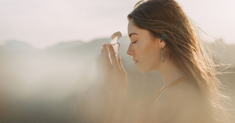 A woman with her eyes closed, holding a clear crystal gently to her forehead in soft morning light